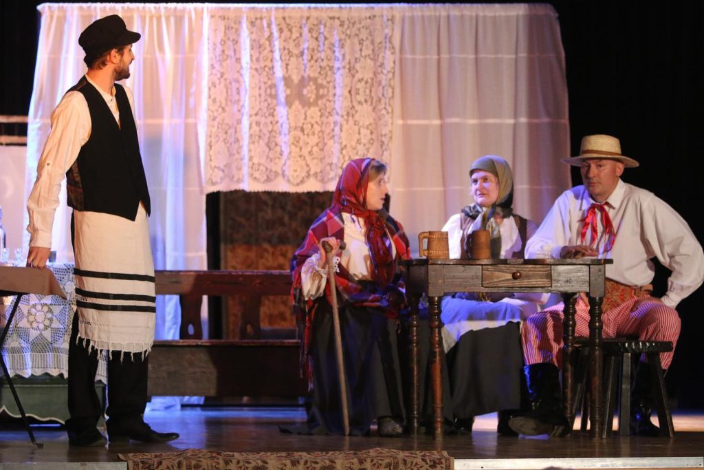 aktorzy podczas spektaklu. po lewej mężczyzna, po prawej przy stole dwie kobiety i mężczyna. Wszyscy ubrani w stylu staropolskim