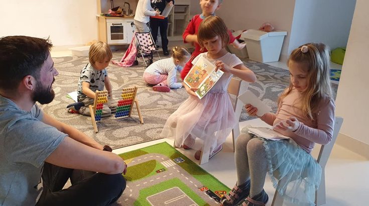 na pierwszym planie mężyczna i dwie małe dziewczynki które trzymają książeczkę i uczą mężczyznę czytac. w tle dzieci bawiące się na dywanie