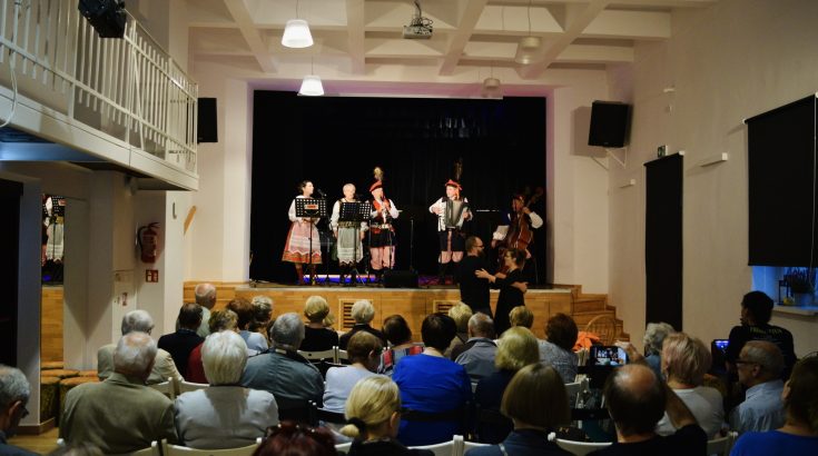 Na pierwszym planie na scenie w trakcie koncertu gra zespól folklorystyczny ubrany w stroje ludowe. tyłem siedzi liczna publiczność