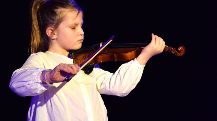 Dziewczynka ubrana w białą bluzkę, stojąca na scenie i grająca za skrzypcach