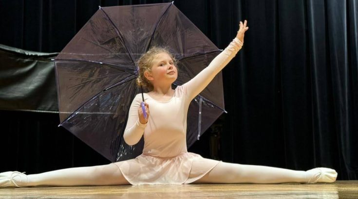 dziewczynka w stroju baletowym w szpagacie. Lewa ręka uniesiona w pozycji baletowej, w prawej ręce parasolka